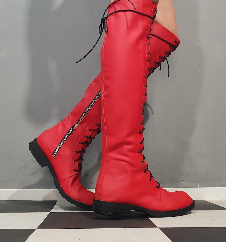 May include: Bright red knee-high boots with black laces and zippers. The boots have a flat sole and a smooth, shiny finish. The boots are shown on a black and white checkered floor.