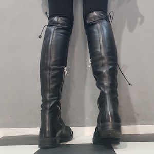 May include: Black leather over-the-knee boots with side zippers and tie closures at the top. The boots have a low heel and are worn with black leggings. The background is a gray wall and a black and white checkered floor.