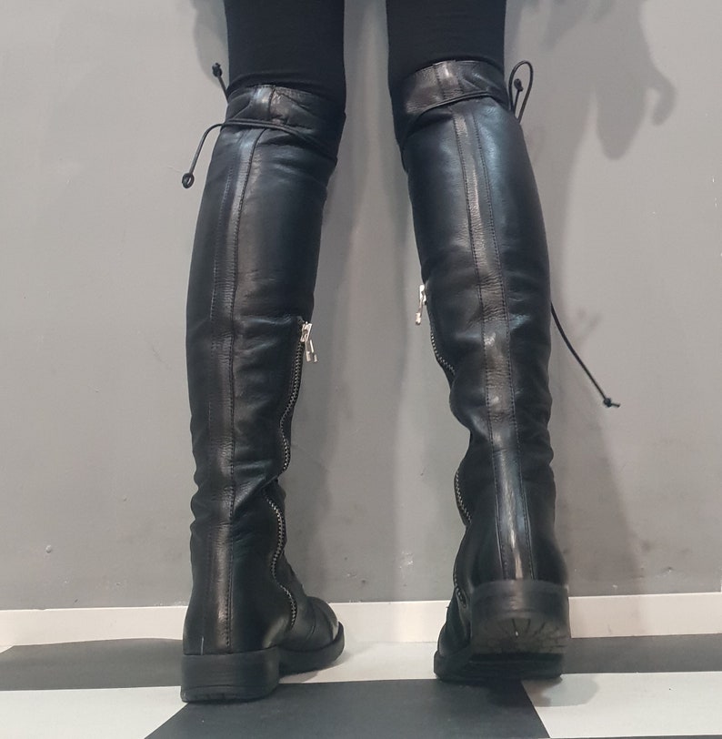 May include: Black leather over-the-knee boots with side zippers and tie closures at the top. The boots have a low heel and are worn with black leggings. The background is a gray wall and a black and white checkered floor.