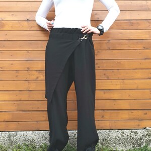 May include: Black wrap-style pants with a wide leg design. The pants feature a unique wrap detail at the front, secured with a gold-colored metal clasp. The model is wearing a white long-sleeve shirt and black shoes. The background is a wooden wall.