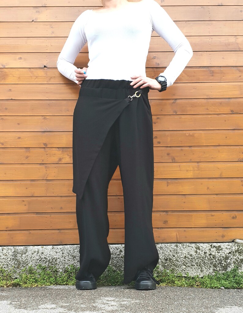 May include: Black wrap-style pants with a wide leg design. The pants feature a unique wrap detail at the front, secured with a gold-colored metal clasp. The model is wearing a white long-sleeve shirt and black shoes. The background is a wooden wall.