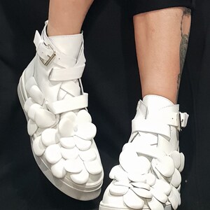 May include: White high-top sneakers with a unique design featuring 3D flower-shaped embellishments. The shoes have a buckle closure and a white sole.