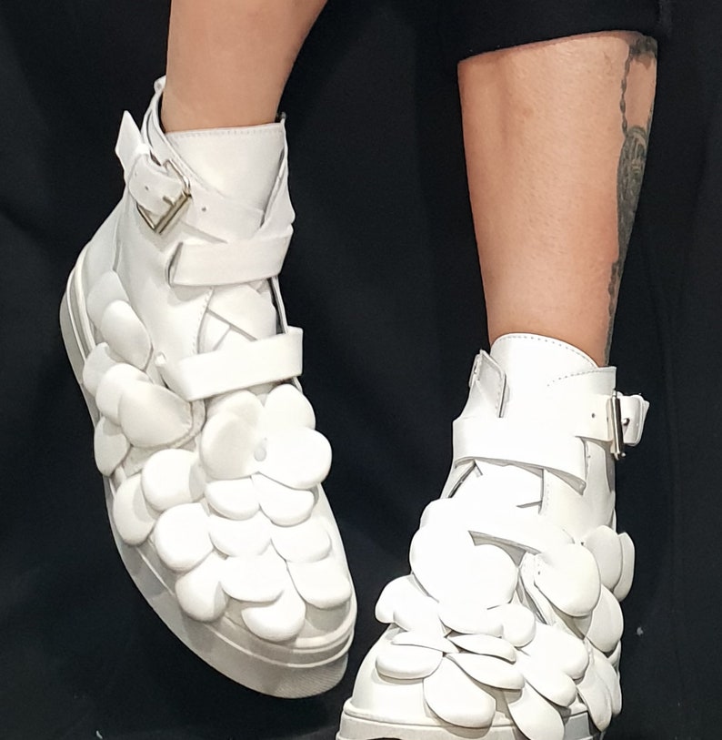 May include: White high-top sneakers with a unique design featuring 3D flower-shaped embellishments. The shoes have a buckle closure and a white sole.