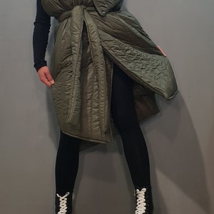 May include: A long, olive green quilted vest with a large collar and a tie belt. The vest is worn over black leggings and black platform boots with white laces.