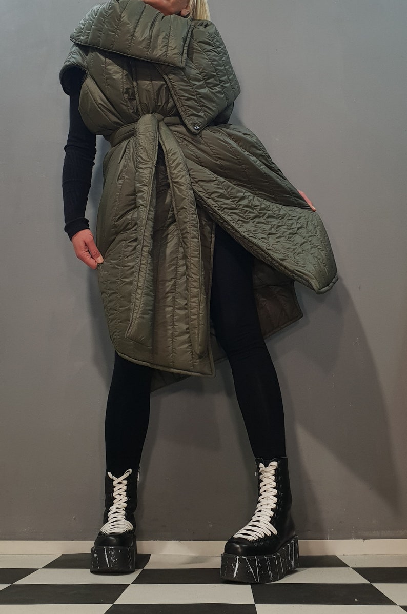 May include: A long, olive green quilted vest with a large collar and a tie belt. The vest is worn over black leggings and black platform boots with white laces.