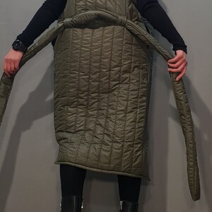 May include: A long, quilted, olive green coat with a high collar and a belt. The coat has a unique design with the sleeves ending at the shoulders. The model is wearing black trousers and black platform boots with white accents.