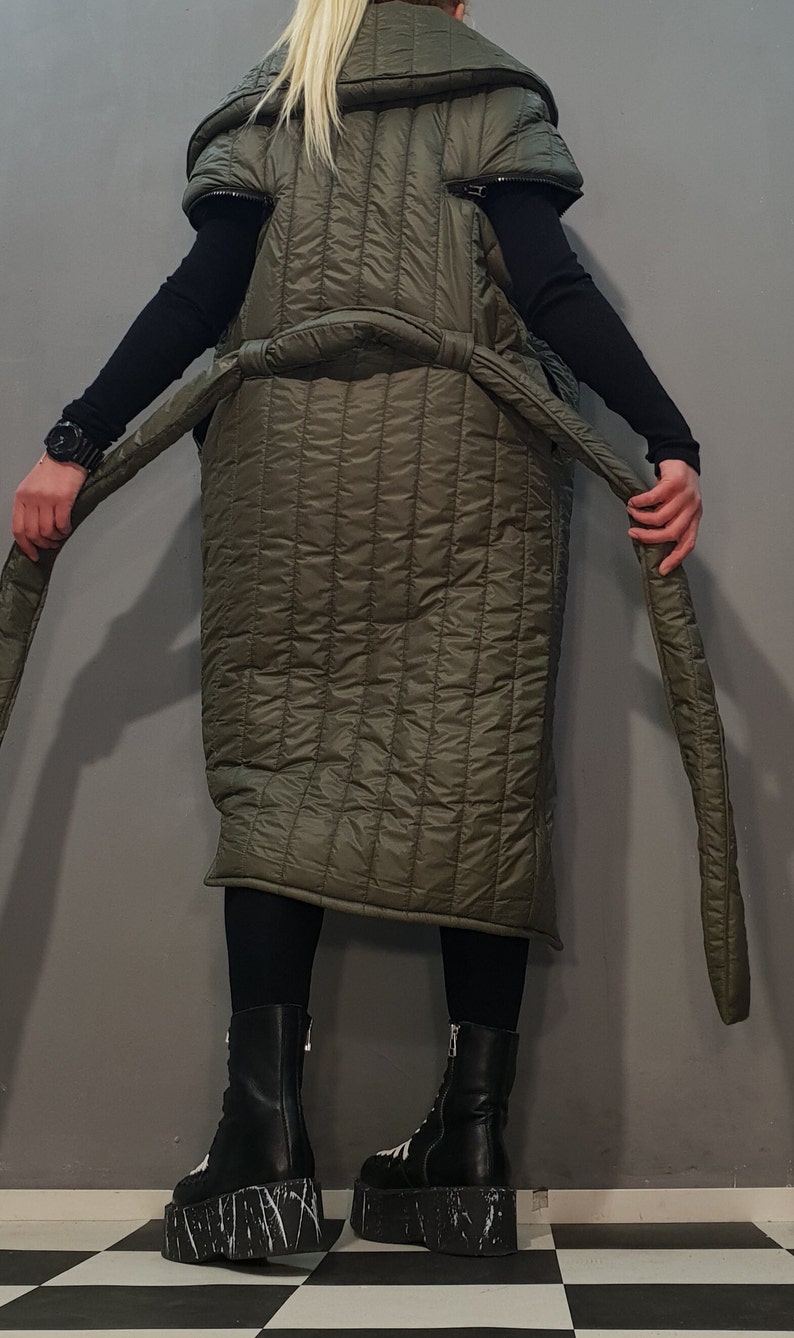 May include: A long, quilted, olive green coat with a high collar and a belt. The coat has a unique design with the sleeves ending at the shoulders. The model is wearing black trousers and black platform boots with white accents.