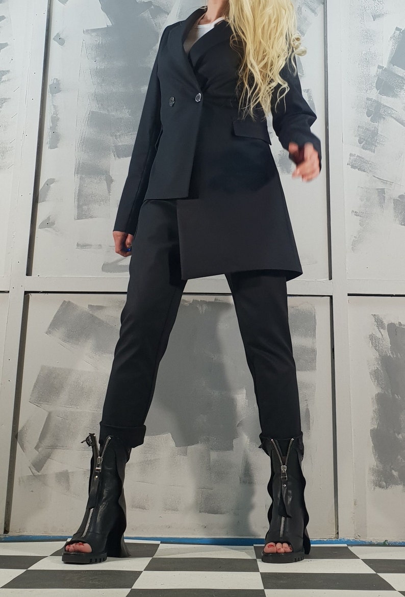 May include: A black blazer with a double-breasted closure and a long, asymmetrical hemline. The blazer is paired with black pants and black heeled boots with zippers.