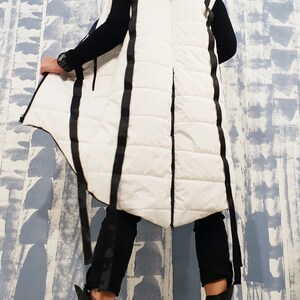 May include: White hooded puffer vest with black straps and a long, asymmetrical hemline. The vest is quilted and has a zipper closure.