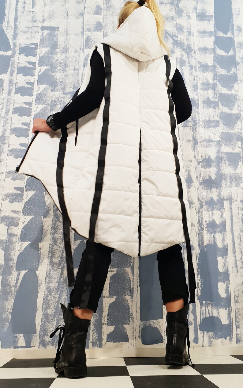 May include: White hooded puffer vest with black straps and a long, asymmetrical hemline. The vest is quilted and has a zipper closure.