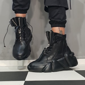 May include: Black leather high-top boots with a unique, geometric design on the soles. The boots feature a front zipper, laces, and a padded tongue. The footwear is worn with black pants.