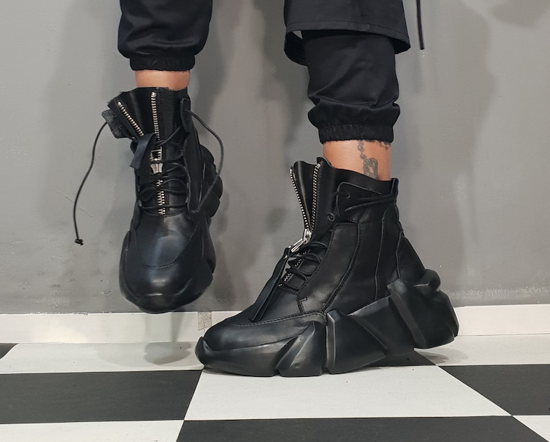May include: Black leather high-top boots with a unique, geometric design on the soles. The boots feature a front zipper, laces, and a padded tongue. The footwear is worn with black pants.
