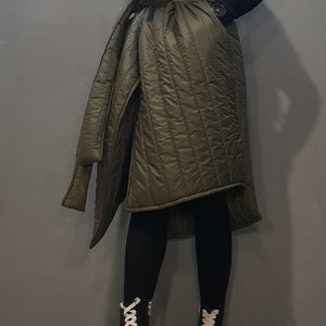 May include: A long, olive green quilted vest with a large collar and a belt. The vest is worn over black leggings and black platform boots with white laces.