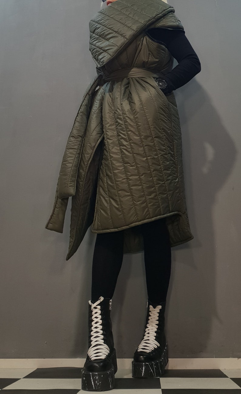 May include: A long, olive green quilted vest with a large collar and a belt. The vest is worn over black leggings and black platform boots with white laces.