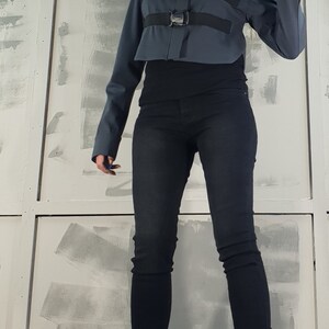 May include: A woman wearing a dark gray cropped jacket with black straps and buckles, black jeans, and black lace-up boots. The jacket has a high collar and long sleeves.