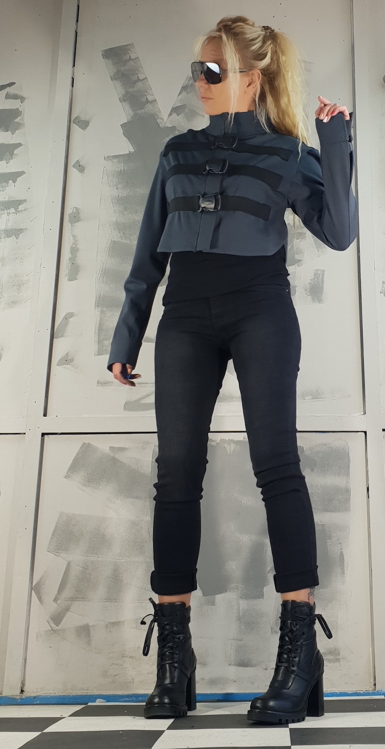 May include: A woman wearing a dark gray cropped jacket with black straps and buckles, black jeans, and black lace-up boots. The jacket has a high collar and long sleeves.