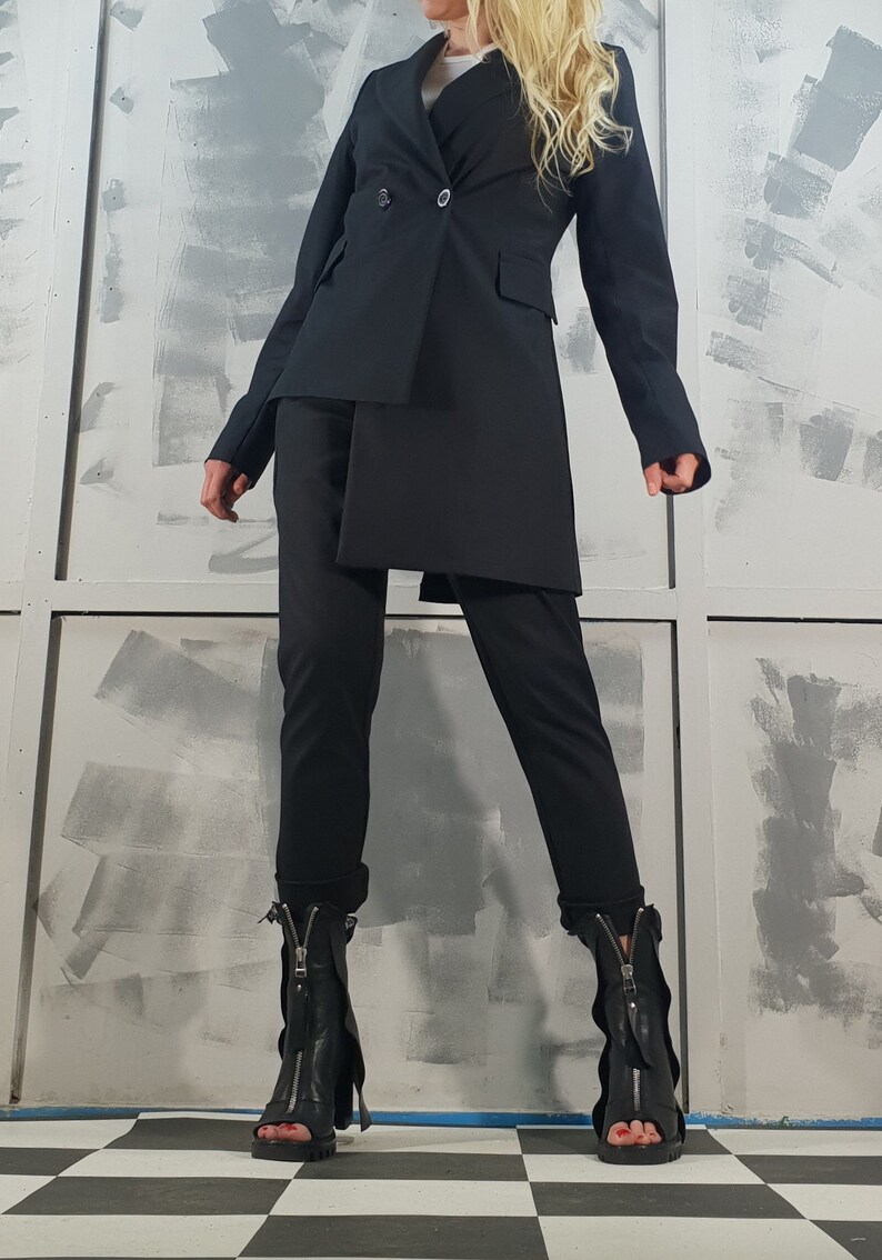 May include: A black blazer with a long, asymmetrical hemline, worn with black pants and black heeled boots with zippers.