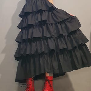 May include: A black sleeveless dress with multiple layers of ruffles. The dress has a zipper closure in the front. The model is wearing red open-toe platform boots.