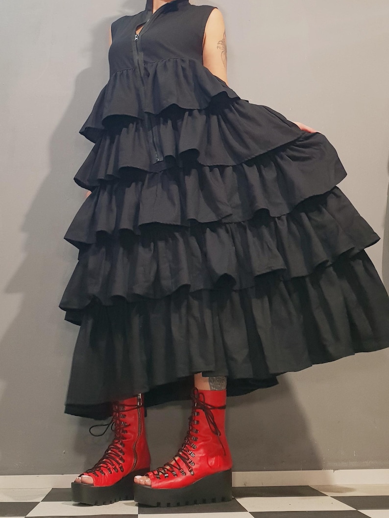 May include: A black sleeveless dress with multiple layers of ruffles. The dress has a zipper closure in the front. The model is wearing red open-toe platform boots.
