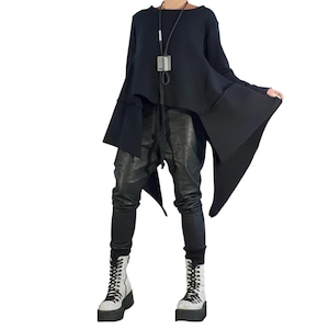 Op de afbeelding: Een zwarte tuniek met lange mouwen en een asymmetrische zoom, gecombineerd met een zwarte broek en witte veterschoenen met plateauzolen. Een lange ketting met een rechthoekige hanger hangt om de hals bij deze donkere kledingcombinatie.