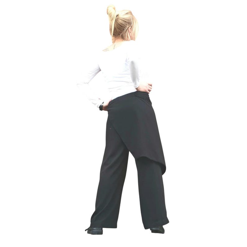 May include: Black wrap-style pants with a wide leg design. The pants feature a unique wrap detail at the waist, creating an asymmetrical look. The model is wearing a white long-sleeve shirt. The pants are made of a soft, flowing fabric.