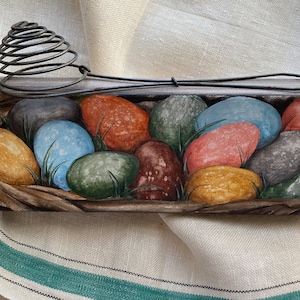 May include: A painting of a nest filled with colorful Easter eggs. The eggs are painted in shades of blue, green, red, yellow, and brown. A vintage wire egg whisk sits on top of the nest.