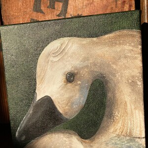 May include: A square canvas painting of a swan's head and neck. The swan has a black beak and a light beige head and neck. The background is a dark green color. The painting is on a wooden surface.