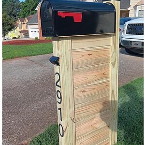 May include: A black metal mailbox with a red flag mounted on a vertical wooden post. The post features horizontal wood paneling and the black house numbers 2910 displayed vertically on the side. A small black solar light is attached to the post.