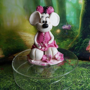 Könnte beinhalten: Eine weiße Mausfigur, die ein rosa Kleid trägt und rosa Blumen hält. Die Figur steht auf einem klaren Glassockel.