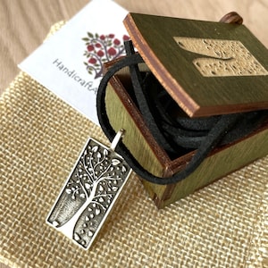 Könnte beinhalten: Ein rechteckiger Silberanhänger mit Baumdesign, an einer schwarzen Kordel, liegt in einer offenen grünen Holzbox. Eine kleine Karte mit dem Text "HandicraftArt" und einem Baumlogo ist sichtbar. Die Box steht auf einer Jutefläche.