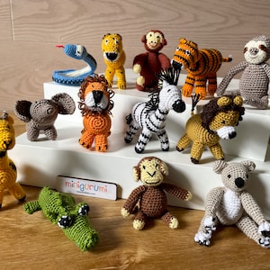 May include: A collection of 12 crocheted animal toys, including a giraffe, elephant, lion, zebra, monkey, tiger, sloth, snake, crocodile, and koala. The toys are all different colours and are arranged on a wooden surface. The text "miniguru mi" is visible on a wooden sign.