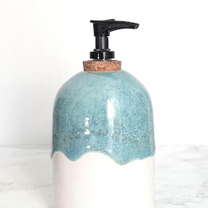 May include: A two-tone ceramic soap dispenser. The top half is a speckled teal glaze, the bottom half is white. It features a black pump and a cork base. A decorative bathroom accessory.