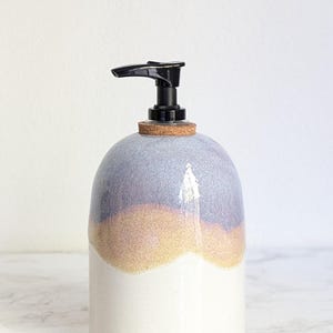 May include: A ceramic soap dispenser with a rounded body featuring a three-tone glaze in lavender, soft orange, and white. The top includes a cork collar and a black plastic pump mechanism for dispensing liquid soap or lotion.