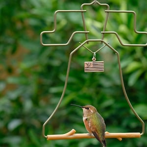 May include: A hummingbird feeder shaped like a bell with a small American flag charm hanging from it. A hummingbird is perched on a wooden perch.