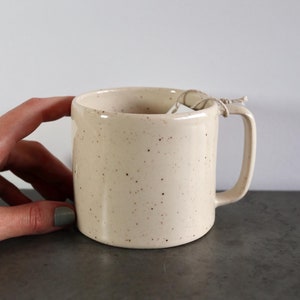 Puede incluir: Una taza de cerámica blanca moteada con asa. La taza está sobre una superficie gris.