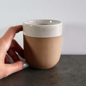 Puede incluir: Una taza de cerámica con cuerpo marrón claro y borde blanco. La taza es ligeramente redondeada y tiene un acabado mate.