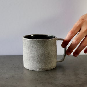 May include: A grey ceramic mug with a black interior and a handle. The mug has a speckled texture.