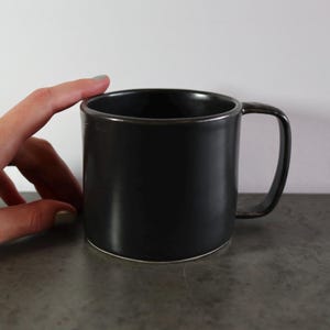 Puede incluir: Una taza de cerámica negra con forma redondeada y asa. La taza está sobre una superficie gris.