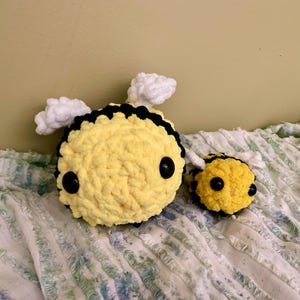 以下が含まれることがあります： 2つの編みぐるみの蜂のぬいぐるみ。大きな蜂は黄色で、黒い縞模様、黒い目、白い羽があります。小さな蜂も同じデザインです。ぬいぐるみは白と青の模様の表面にあります。