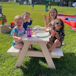 May include: Four children sit at a white picnic table eating pizza. The table has a white wooden top and white wooden legs. The children are sitting on the benches of the table. There is a green grassy area behind the table.