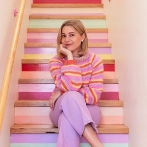 Op de afbeelding: Een vrouw in een gestreepte trui en lavendelkleurige broek zit op een kleurrijke trap. De trap heeft afwisselend roze, gele, groene en paarse treden.