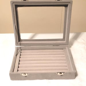 May include: A rectangular gray jewelry display case with a clear glass lid and a velvet-lined interior. The box features seven horizontal padded slots for organizing rings or small items, secured by two silver-toned metal latches on the front edge.