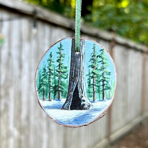 May include: Hand-painted wooden ornament featuring a forest scene with a large tree and snow. The round ornament is painted with shades of green, blue, and brown, and is suspended by a green ribbon. The ornament is approximately 3 inches in diameter.