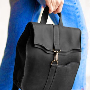 Pode incluir: Uma mochila de couro preto fosco com al&ccedil;a superior, aba com uma tira vertical central e fecho de mosquet&atilde;o met&aacute;lico. A bolsa inclui um bolso frontal e al&ccedil;as de ombro ajust&aacute;veis com costuras vis&iacute;veis em toda a sua estrutura.