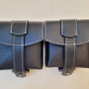 Pu&ograve; includere: Due borse in pelle nera con cuciture bianche e chiusura a fibbia. Le borse sono collegate tra loro.