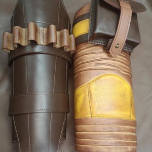 Może przedstawiać: Para brązowych sk&oacute;rzanych ochraniaczy na nogi z ż&oacute;łto-brązowym paskowanym wzorem. Jeden ochraniacz ma małą brązową sk&oacute;rzaną kieszeń z paskiem.