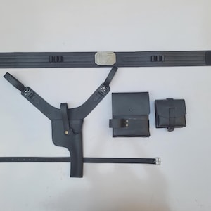 May include: A black leather holster set with a belt, shoulder straps, and two pouches. The belt has a silver buckle with a design on it.