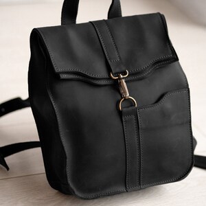 Pode incluir: Uma mochila de couro preto fosco com al&ccedil;a superior, al&ccedil;as de ombro ajust&aacute;veis e aba. A aba &eacute; fixada com um fecho de mosquet&atilde;o de metal em tom de lat&atilde;o preso a uma tira de couro vertical no painel frontal da mochila.