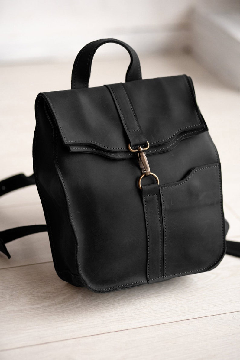 Pode incluir: Uma mochila de couro preto fosco com al&ccedil;a superior, al&ccedil;as de ombro ajust&aacute;veis e aba. A aba &eacute; fixada com um fecho de mosquet&atilde;o de metal em tom de lat&atilde;o preso a uma tira de couro vertical no painel frontal da mochila.