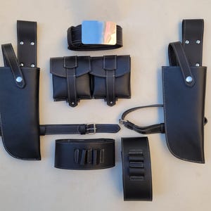 Pu&ograve; includere: Un set di fondine in pelle nera con cuciture bianche, che comprende una cintura, due doppie tasche e due tasche singole. Il set include anche una fibbia per cintura in metallo argentato.